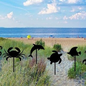 May include: A set of eight black metal silhouette garden stakes featuring various sea creatures, including a seahorse, octopus, crab, dolphin, turtle, fish, and mermaid. The stakes are displayed in a grassy area with a beach and ocean in the background.
