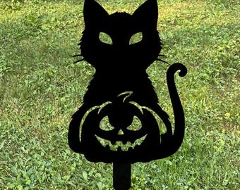 Może przedstawiać: Czarny metalowy palik ogrodowy na Halloween z sylwetką kota siedzącego nad wyrzeźbioną dynią. Kot ma spiczaste uszy i zakrzywiony ogon. Dynia ma wyrzeźbioną twarz z trójkątnymi oczami i uśmiechem.