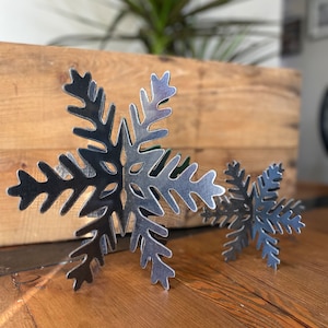 May include: Two metal snowflakes, one larger than the other, are displayed on a wooden surface. The snowflakes are a silvery color and have a detailed design.