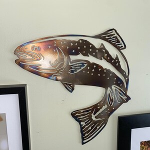 Puede incluir: Una escultura de pared de metal de una trucha con un acabado marr&oacute;n, azul y plateado. El pez est&aacute; en una pose de salto con la cola levantada.