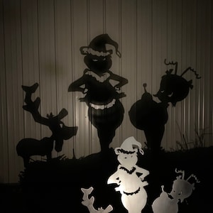 Peut inclure: Décorations de jardin de Noël en silhouette métallique représentant le Grinch et des rennes. Les personnages du Grinch portent des chapeaux de Père Noël et ont une expression espiègle. Les rennes ont de grands bois. Les personnages sont placés sur un fond sombre.