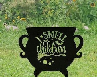 Pode incluir: Arte de jardim de caldeirão de metal preto com o texto "I smell children". O caldeirão tem duas alças e três pernas. O texto inclui um morcego, estrelas e uma vassoura de bruxa. O caldeirão é colocado em um gramado.