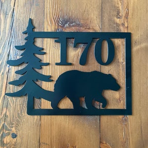 Peut inclure: Plaque de numéro de maison en métal noir avec une silhouette d'un pin et d'un ours. Le numéro 170 est affiché sur la plaque.