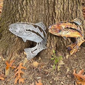 Puede incluir: Dos esculturas de peces de metal, una plateada y otra con p&aacute;tina marr&oacute;n, se exhiben contra un tronco de &aacute;rbol. Los peces est&aacute;n en una pose de salto con las colas levantadas.