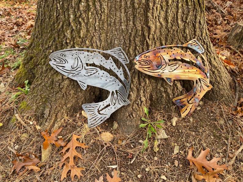 Puede incluir: Dos esculturas de peces de metal, una plateada y otra con p&aacute;tina marr&oacute;n, se exhiben contra un tronco de &aacute;rbol. Los peces est&aacute;n en una pose de salto con las colas levantadas.