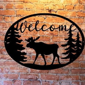 May include: Black metal oval sign with the word "Welcome" and a silhouette of a moose standing in front of two pine trees.