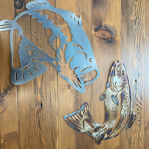 Puede incluir: Arte de pared de metal con dos siluetas de peces. El pez m&aacute;s grande es una lubina con un dise&ntilde;o detallado, mientras que el pez m&aacute;s peque&ntilde;o es una trucha con un dise&ntilde;o m&aacute;s abstracto. Ambos peces est&aacute;n en posici&oacute;n de salto, creando una escena din&aacute;mica y animada.