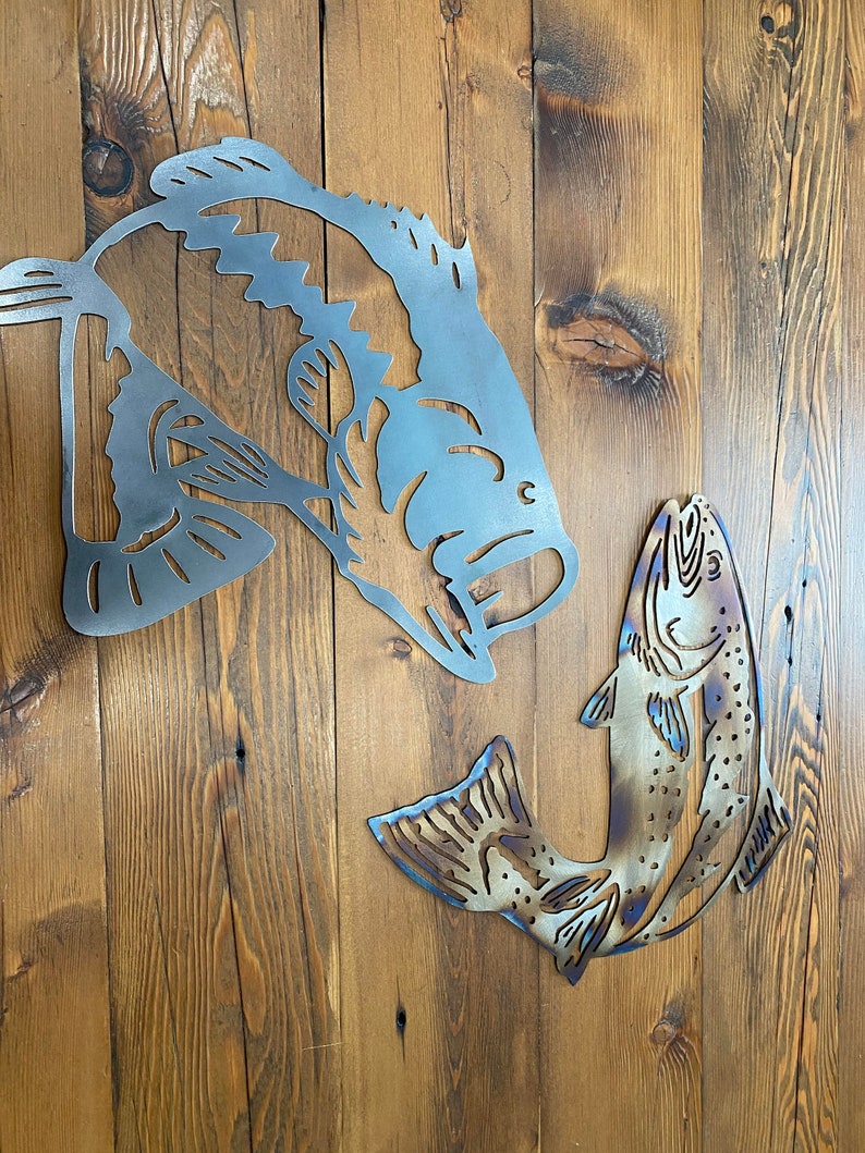 Puede incluir: Arte de pared de metal con dos siluetas de peces. El pez m&aacute;s grande es una lubina con un dise&ntilde;o detallado, mientras que el pez m&aacute;s peque&ntilde;o es una trucha con un dise&ntilde;o m&aacute;s abstracto. Ambos peces est&aacute;n en posici&oacute;n de salto, creando una escena din&aacute;mica y animada.