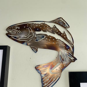 Puede incluir: Una escultura de pared de metal de una trucha marr&oacute;n saltando fuera del agua. La escultura tiene un aspecto r&uacute;stico y envejecido con una p&aacute;tina marr&oacute;n y azul.