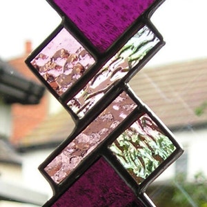 Puede incluir: Un atrapasol de vidrio emplomado con un diseño geométrico que presenta piezas de vidrio morado, rosa y blanco. El atrapasol está colgado de una ventana.