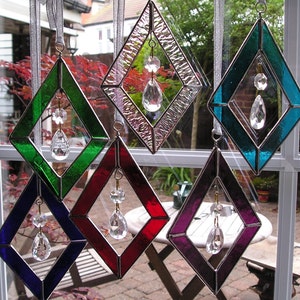 Op de afbeelding: Vijf diamantvormige gebrandschilderde zonvangers met kristallen prisma's die eraan hangen. De zonvangers zijn in verschillende kleuren, waaronder groen, rood, paars, blauw en helder.