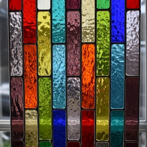 Könnte beinhalten: Ein Buntglasfenster mit einem bunten Ziegelmuster. Das Fenster zeigt eine Vielzahl von Farben, darunter Rot, Orange, Gelb, Grün, Blau und Lila. Das Glas ist strukturiert und hat einen leicht schillernden Glanz.