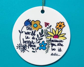 May include: Round white ornament with a gold string, featuring a floral design and the text "the world is a better place because you are in it." The ornament has blue, yellow, and pink flowers.