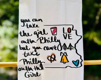 May include: White kitchen towel with a hand-drawn design. The towel features the text "you can take the girl outta Philly but you can't take Philly outta the Girl." The design includes a red drink, the word "LOVE", a bridge, a pretzel, a bell, and a blue heart.