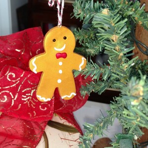 May include: A gingerbread man ornament with white icing and red bow tie hanging on a green artificial Christmas tree.