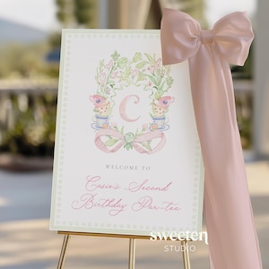 May include: A welcome sign on a gold easel with a pink bow. The sign features a floral wreath design with teacups and the letter "C". Text reads "Welcome to Casie's Second Birthday Par-tea".
