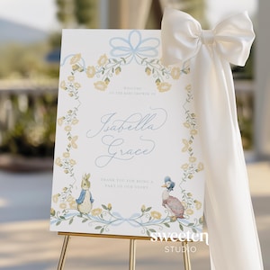 May include: A welcome sign for a baby shower, featuring the name "Isabella Grace" in elegant script. The sign is decorated with floral and ribbon accents in yellow and blue, and includes illustrations of Peter Rabbit and a duck. A large white bow adorns the side.