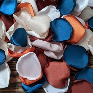 Op de afbeelding: Een verzameling zijden bloemblaadjes in tinten wit, roest, oranje en teal. De bloemblaadjes zijn op een houten oppervlak in een stapel gerangschikt.