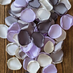 May include: A collection of silk flower petals in shades of purple, white, and grey. The petals are arranged on a wooden surface.