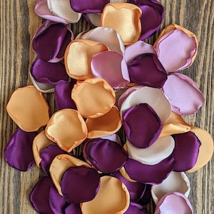 Könnte beinhalten: Ein Haufen Seidenblumenblätter in den Farben Lila, Pfirsich und Orange. Die Blütenblätter sind in einem zufälligen Muster angeordnet und eignen sich perfekt für Hochzeitsdekorationen oder andere besondere Anlässe.
