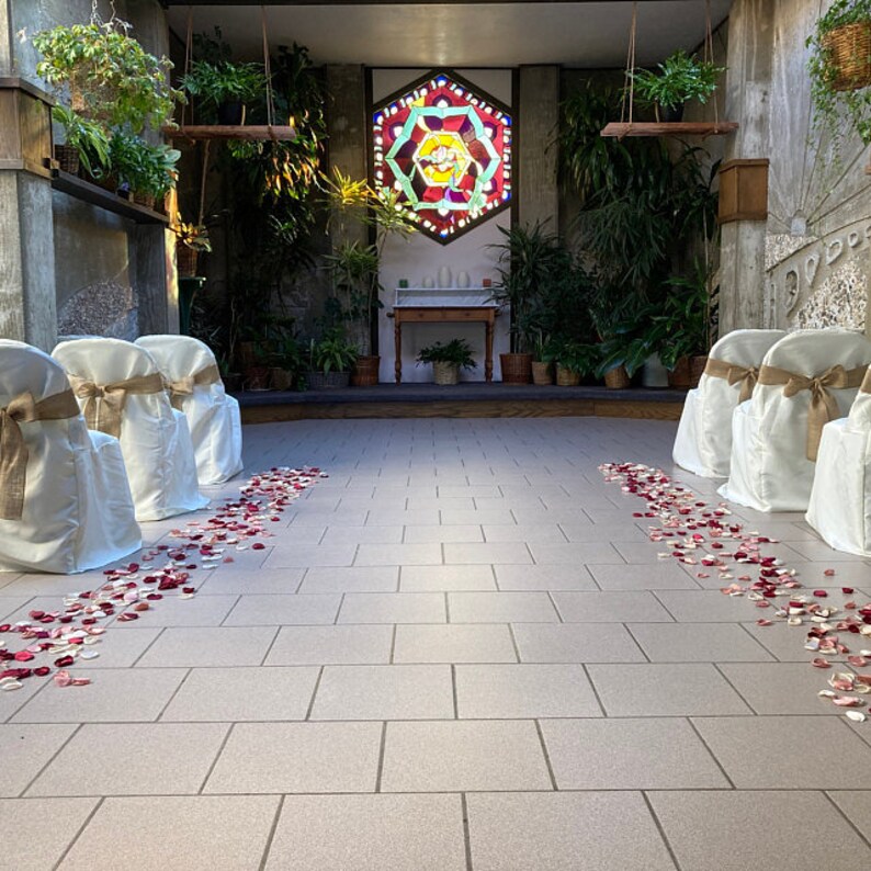 Puede incluir: Un pasillo de ceremonia de boda decorado con p&eacute;talos de rosa, con sillas blancas alineadas a cada lado. Se puede ver una ventana de vidrieras con un dise&ntilde;o geom&eacute;trico en el fondo.
