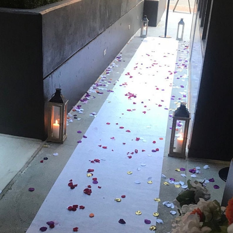 May include: A white aisle runner with scattered rose petals in shades of pink, purple, and red. Two black lanterns with lit candles are placed on either side of the runner.