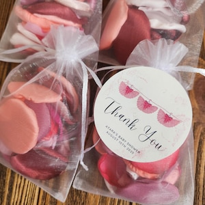 Op de afbeelding: Vier kleine, doorschijnende witte zakjes gevuld met roze en perzik kleurige stoffen bloemblaadjes. Eén zakje heeft een ronde sticker met de tekst "Thank You" en de tekst "Atara's Baby Shower August 15th 2025". De zakjes zijn vastgebonden met witte linten.