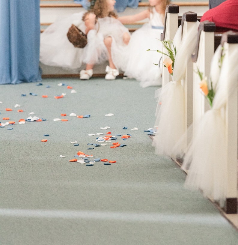 May include: White aisle runner with white tulle bows and orange flowers. Scattered blue, white, and orange flower petals on the floor.