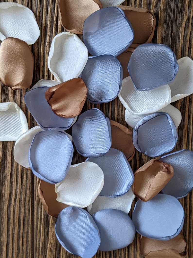 Puede incluir: Una colecci&oacute;n de p&eacute;talos de flores de tela en tonos de blanco, azul claro y marr&oacute;n. Los p&eacute;talos est&aacute;n dispuestos sobre una superficie de madera.