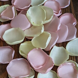 May include: A pile of light pink and pale yellow satin rose petals. The petals are arranged in a random pattern on a wooden surface.