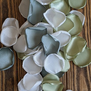 May include: A collection of white, sage green, and gray silk flower petals. The petals are arranged in a pile on a wooden surface.