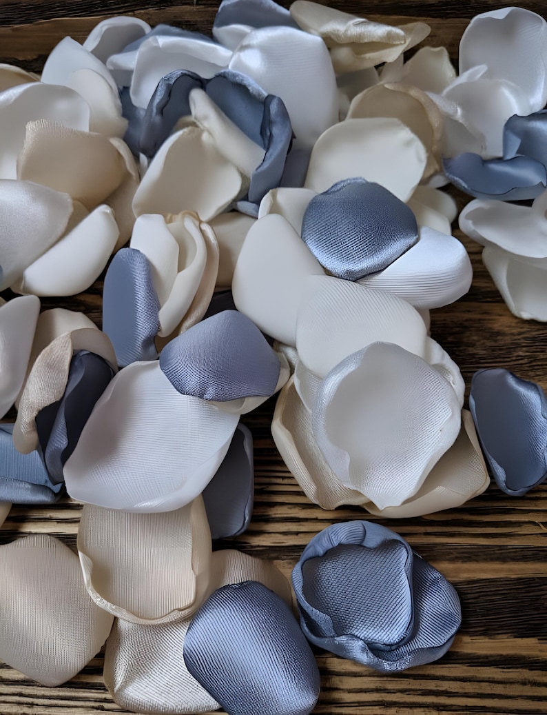 May include: A pile of faux silk rose petals in ivory, white, and light blue. The petals are arranged in a random pattern on a wooden surface.