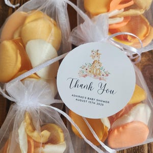 Op de afbeelding: Close-up van verschillende kleine, witte organza zakjes gevuld met gele, oranje en crèmekleurige bloemblaadjes. Elk zakje is vastgebonden met een wit lint en heeft een rond label met "Thank You", een giraffenillustratie en details van een babyshower.