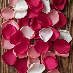 Op de afbeelding: Assortiment van zijden rozenblaadjes in de kleuren wit, roze en fuchsia. De blaadjes zijn verspreid over een houten oppervlak.