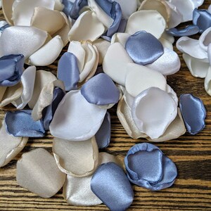 May include: A pile of ivory, white, and dusty blue satin flower petals. The petals are scattered on a wooden surface.