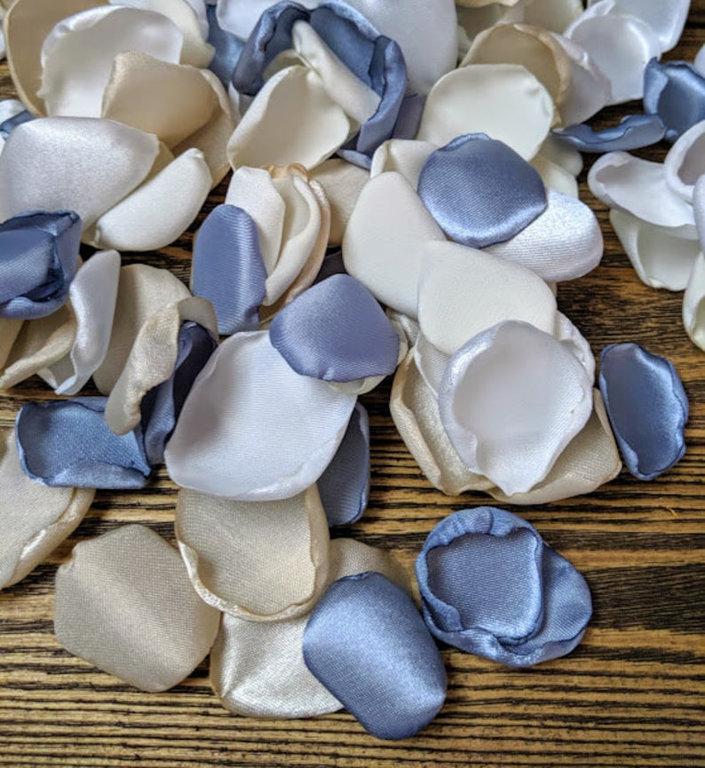 May include: A pile of ivory, white, and dusty blue satin flower petals. The petals are scattered on a wooden surface.