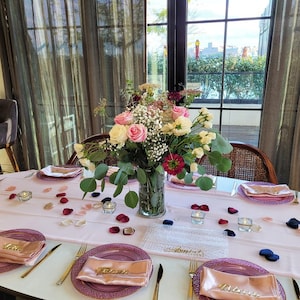 May include: A pink table setting with pink and purple plates, gold silverware, and pink napkins. The table is decorated with pink and red rose petals and small glass candle holders. The centerpiece is a bouquet of pink and white roses, baby's breath, and greenery in a clear glass vase. The table is set for a special occasion, such as a wedding or bridal shower.