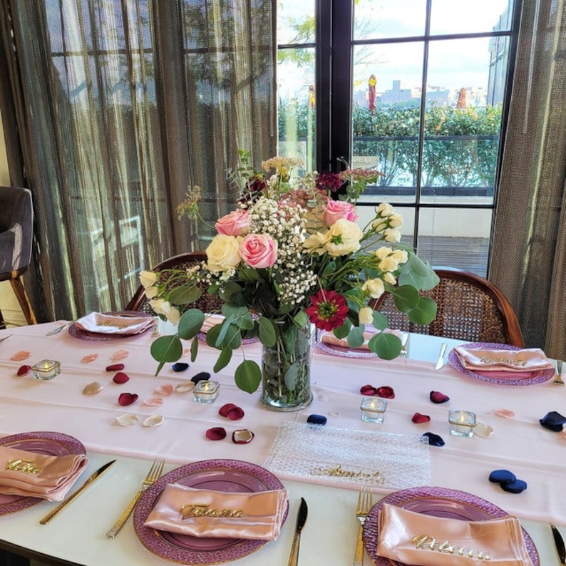 May include: A pink table setting with pink and purple plates, gold silverware, and pink napkins. The table is decorated with pink and red rose petals and small glass candle holders. The centerpiece is a bouquet of pink and white roses, baby's breath, and greenery in a clear glass vase. The table is set for a special occasion, such as a wedding or bridal shower.