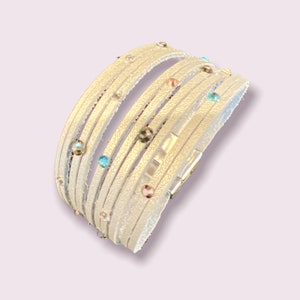 Puede incluir: Un brazalete de cuero blanco con detalles de cristal de colores. El brazalete tiene un cierre plateado.