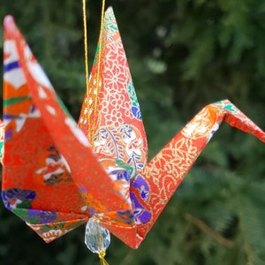May include: A colorful origami crane ornament with a gold string for hanging. The crane is made of patterned paper in shades of red, orange, white, green, and blue. A clear bead hangs below the crane.