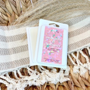 May include: A white package of wax melts labeled "Mells orchards & botanics" with a pink and confetti design. The scent includes apple, citrus, sugar, violet, peach, and musk. The net weight is 2.5 oz.