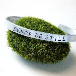 May include: Silver bracelet with the words "Peace Be Still" engraved on it. The bracelet is resting on a bed of green moss.