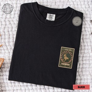 Puede incluir: Camiseta negra con un parche del signo zodiacal Capricornio. El parche presenta una ilustraci&oacute;n de cabra dentro de un dise&ntilde;o circular, con el texto "CAPRICORN" y "DEC 22-JAN 19". La camiseta tiene cuello redondo y mangas cortas.