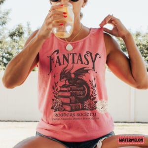 May include: A pink tank top with a black graphic of a dragon sitting on a stack of books. The graphic also includes the text "Fantasy Readers Society" and "Castles, Dragons, Wizards, Heroic Quests".