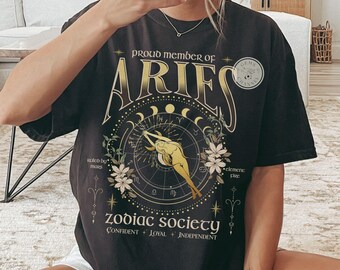 May include: Black t-shirt with a gold graphic of an astrological chart and the text "Proud Member of Aries Zodiac Society". The graphic includes a stylized figure of a person with the text "Confident + Loyal + Independent".