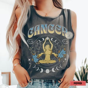 May include: Dark grey vest top featuring a Cancer zodiac design. The graphic includes the word "CANCER" in blue and yellow, a seated figure, and celestial elements. Blue flowers and crescent moons surround the central image. The vest top has a round neckline and sleeveless design.