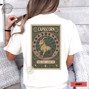 Puede incluir: Camiseta de color marfil con un dise&ntilde;o del signo zodiacal de Capricornio. El gr&aacute;fico incluye un h&iacute;brido cabra-pez, la palabra "CAPRICORN" y las fechas "DEC 22 - JAN 19". El dise&ntilde;o est&aacute; enmarcado con ilustraciones de sol y luna.