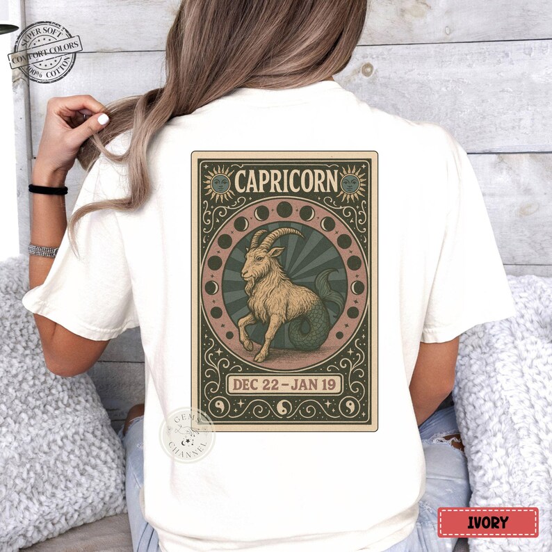 Puede incluir: Camiseta de color marfil con un dise&ntilde;o del signo zodiacal de Capricornio. El gr&aacute;fico incluye un h&iacute;brido cabra-pez, la palabra "CAPRICORN" y las fechas "DEC 22 - JAN 19". El dise&ntilde;o est&aacute; enmarcado con ilustraciones de sol y luna.