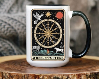 Puede incluir: Taza de café de cerámica blanca con asa negra. La taza presenta un diseño de carta de tarot con fondo negro y una rueda de la fortuna dorada. La carta también presenta un sol, una luna, un pájaro, un león y un perro.
