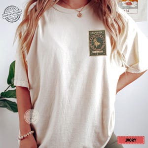 Puede incluir: Camiseta color marfil con un gr&aacute;fico del zodiaco Capricornio en la parte delantera. El gr&aacute;fico presenta una ilustraci&oacute;n de cabra dentro de un marco decorativo. La camiseta tiene un corte holgado y es de algod&oacute;n suave.